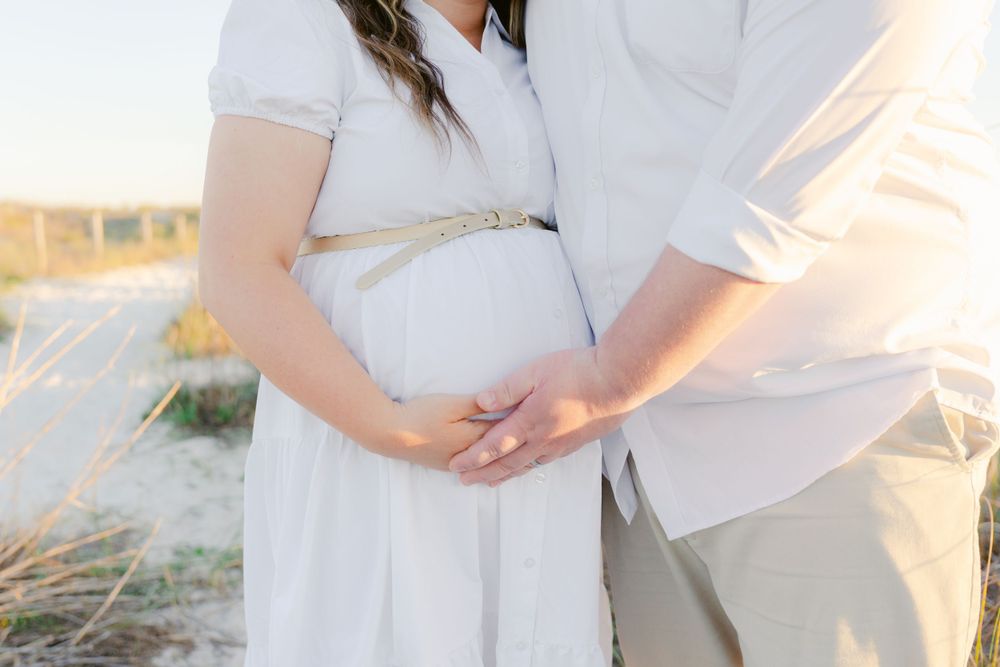 Close-up of couple's hands on pregnant belly against beach backdrop.