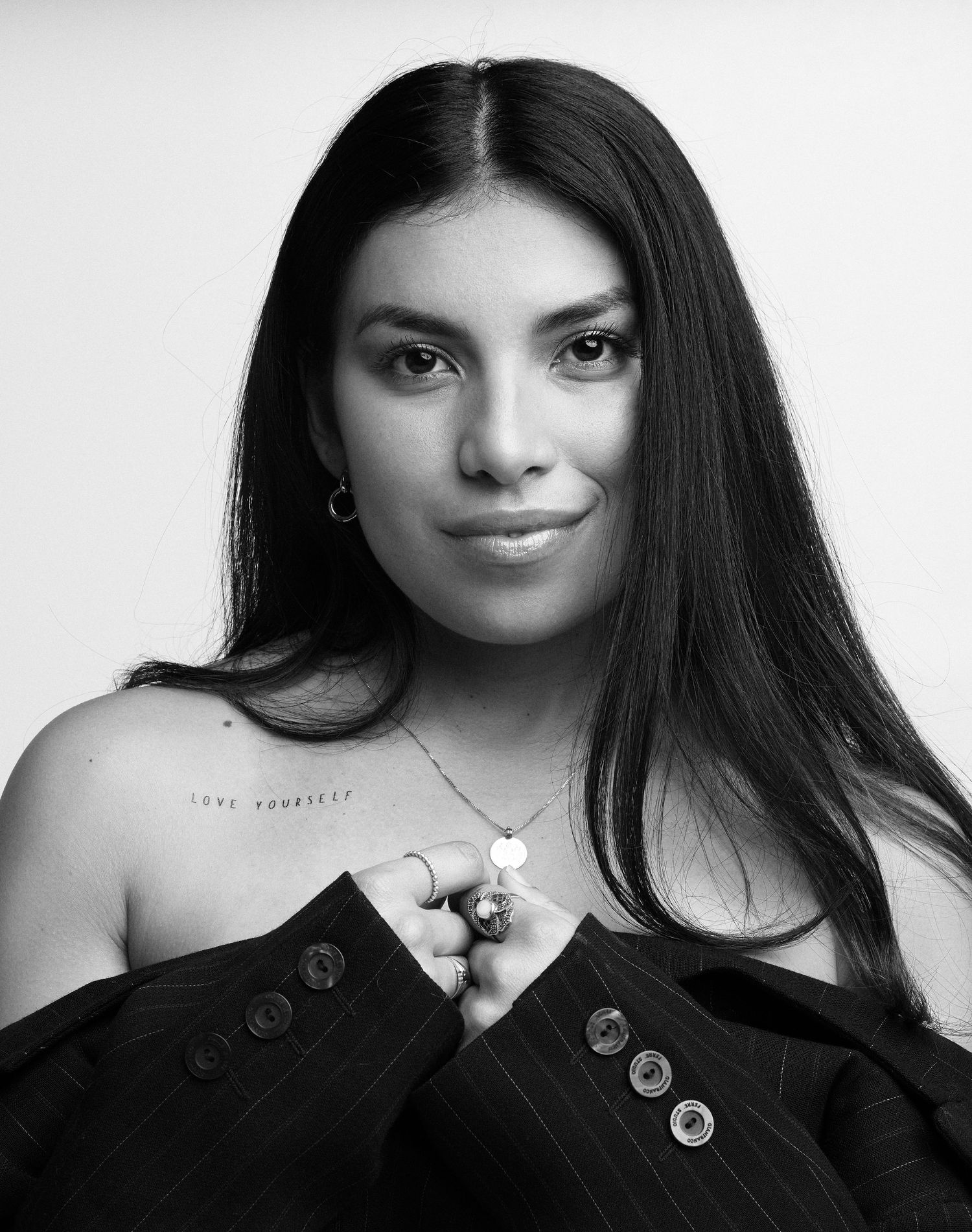 Black and white portrait of a young woman with long dark hair, wearing a blazer and delicate necklace, with a shoulder tattoo.