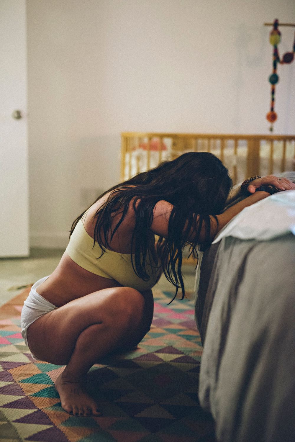 Person in athletic wear crouches on a colorful geometric rug beside a bed in a bedroom setting.