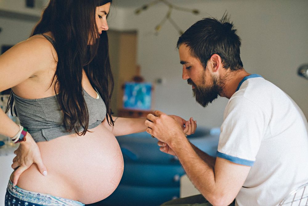 Expectant couple sharing an intimate moment as they admire her pregnant belly in a bedroom setting.