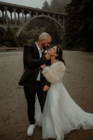 A romantic wedding photo captured on a moody beach with an arched concrete bridge in the misty background.