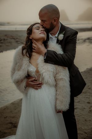 Romantic beach wedding photo of couple embracing in moody lighting with bride wearing white dress and fur wrap.