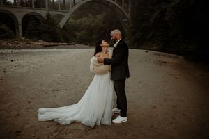 Intimate wedding portrait of couple beneath stone bridge with bride in flowing white gown and groom in black suit.