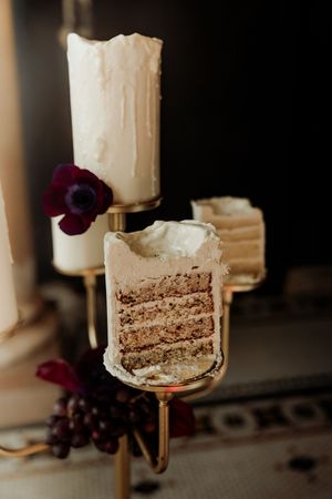 Elegant multi-layer cake slice on gold stand with white frosting, dark floral accents and grapes in moody setting.