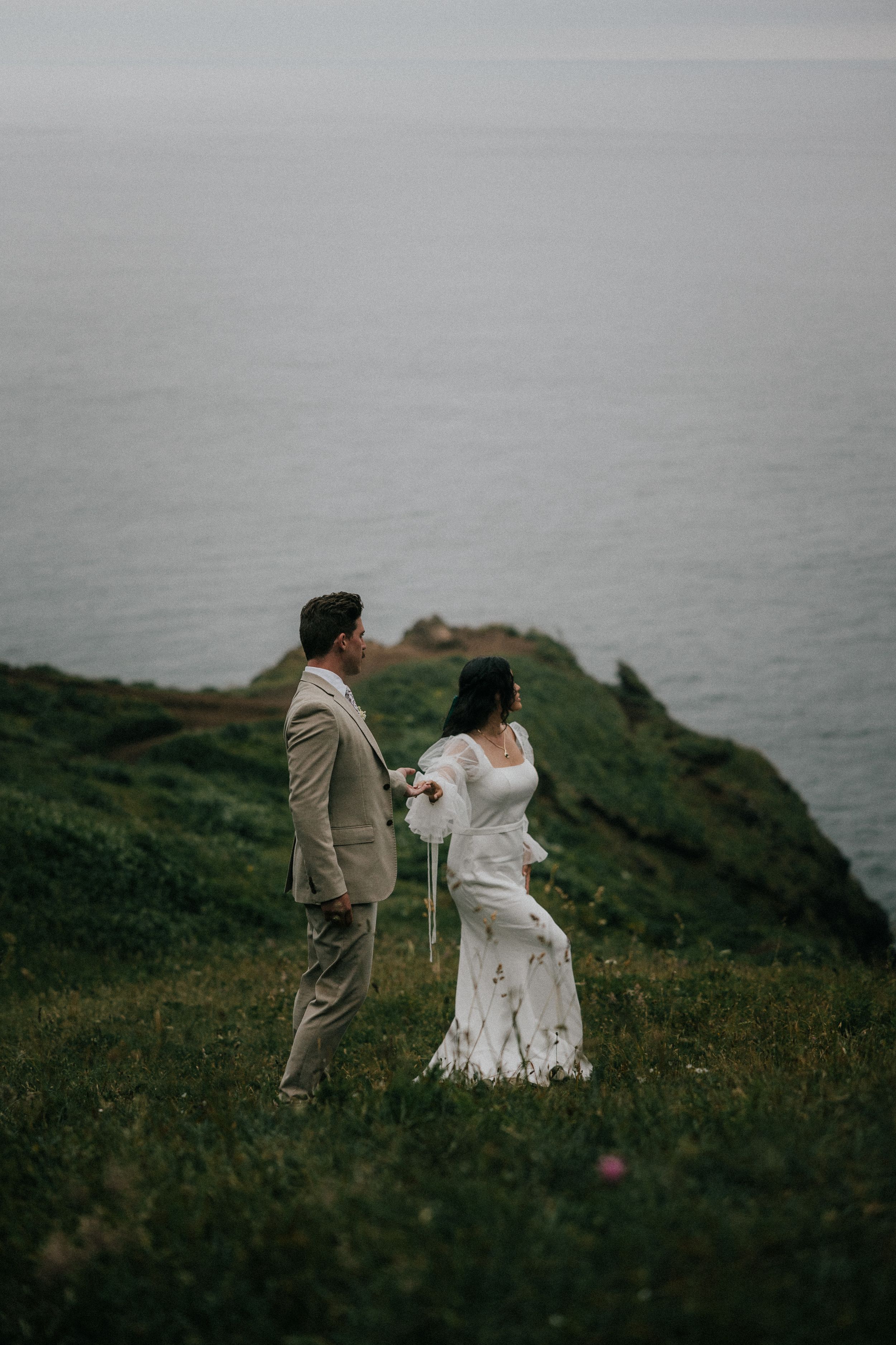 A couple in wedding attire walks along a misty seaside cliff with dramatic coastal views in the background.
