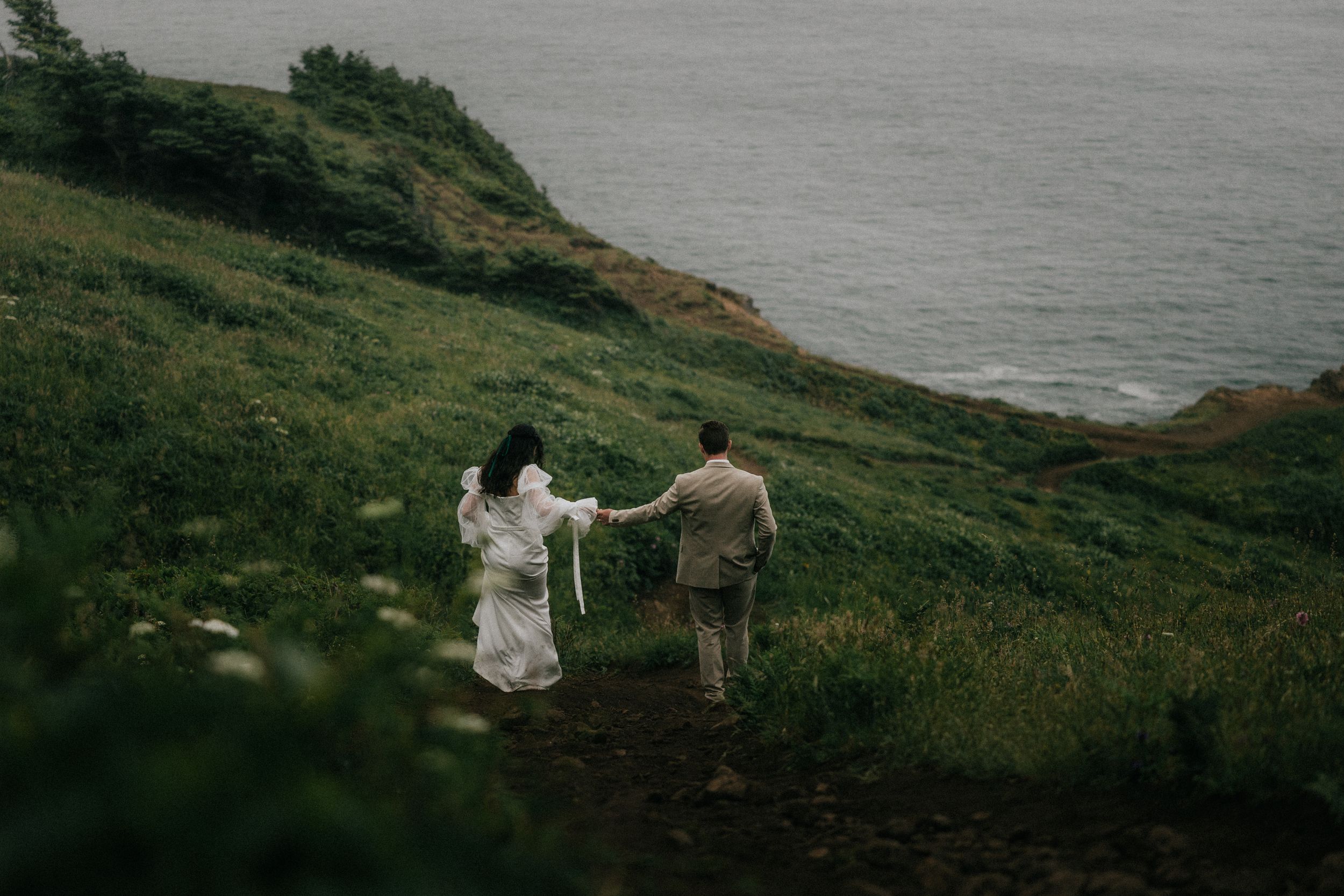 A couple walks hand in hand along a misty coastal cliff trail overlooking the ocean.