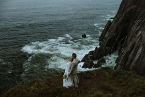 Figure in white dress stands at edge of dramatic coastal cliff overlooking stormy ocean waves crashing below.
