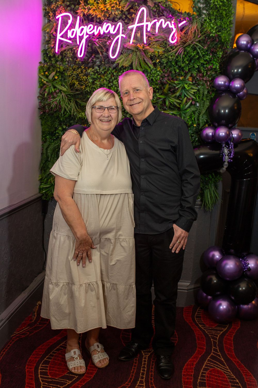 Two people pose together in front of a neon pink 'Ridgeway Arms' sign with purple balloon decor and greenery backdrop.