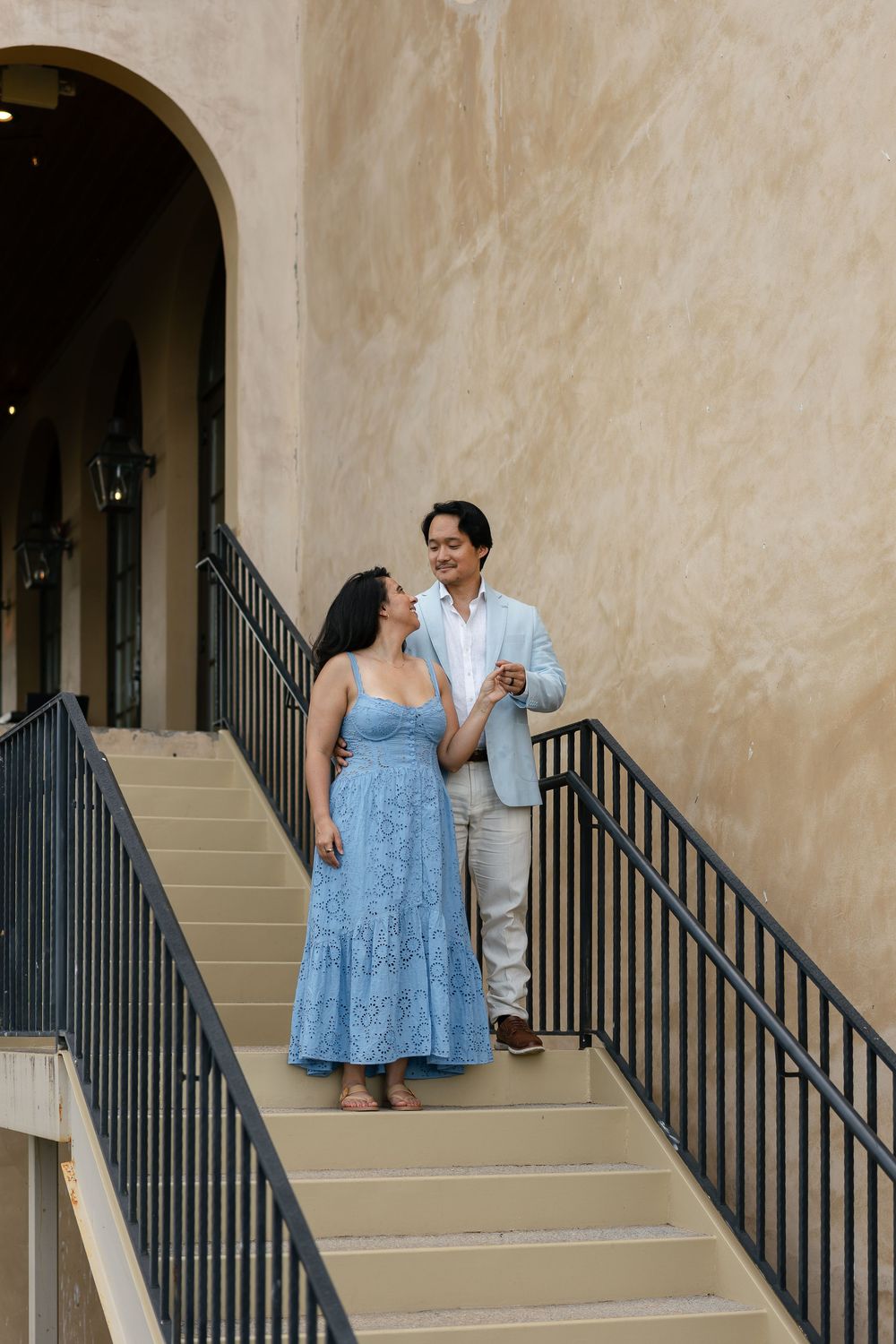 A couple in casual attire stands on concrete stairs with a beige stucco wall and black railing in the background.