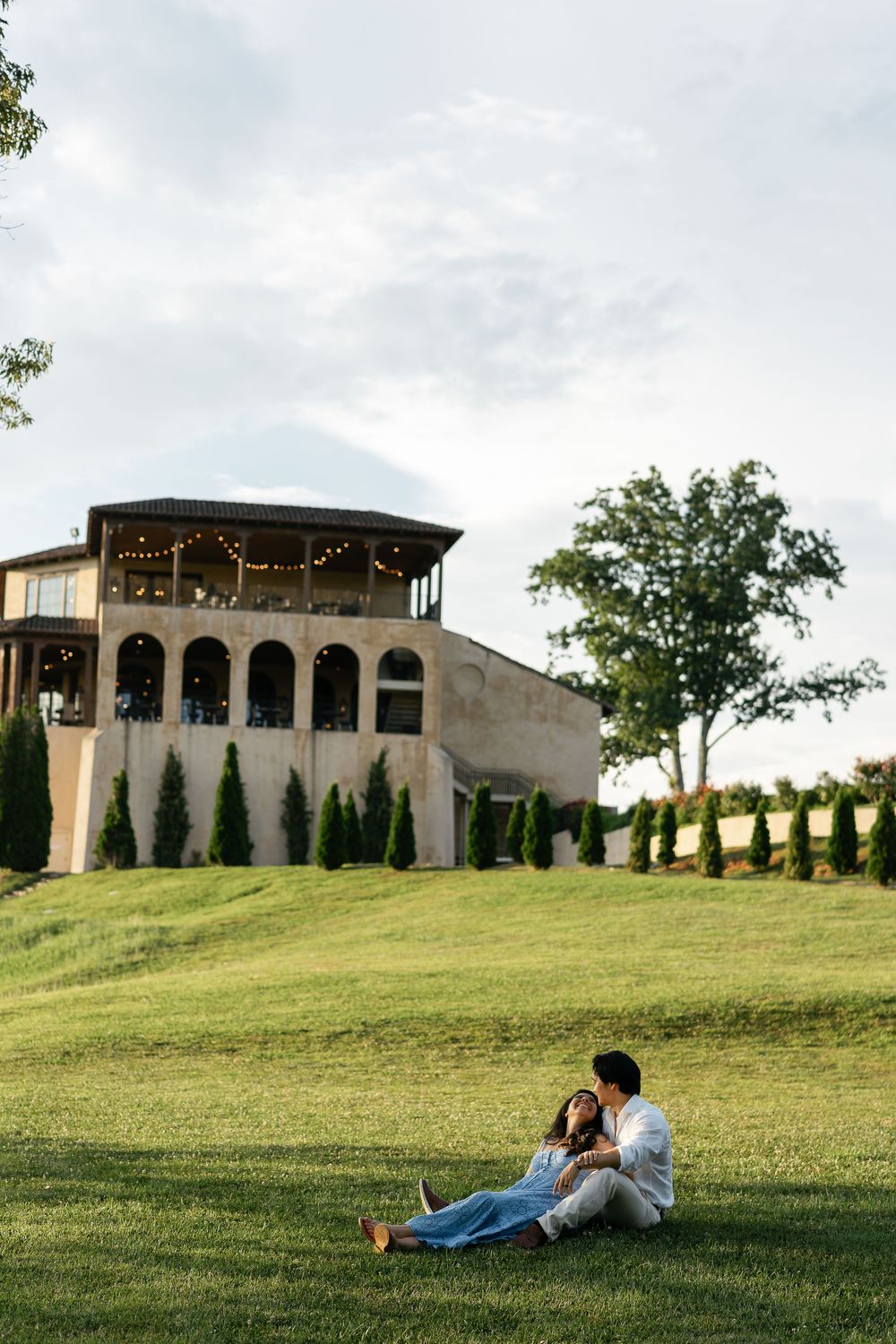 A couple relaxes on a grassy hill overlooking an elegant Mediterranean-style venue decorated with string lights at dusk.