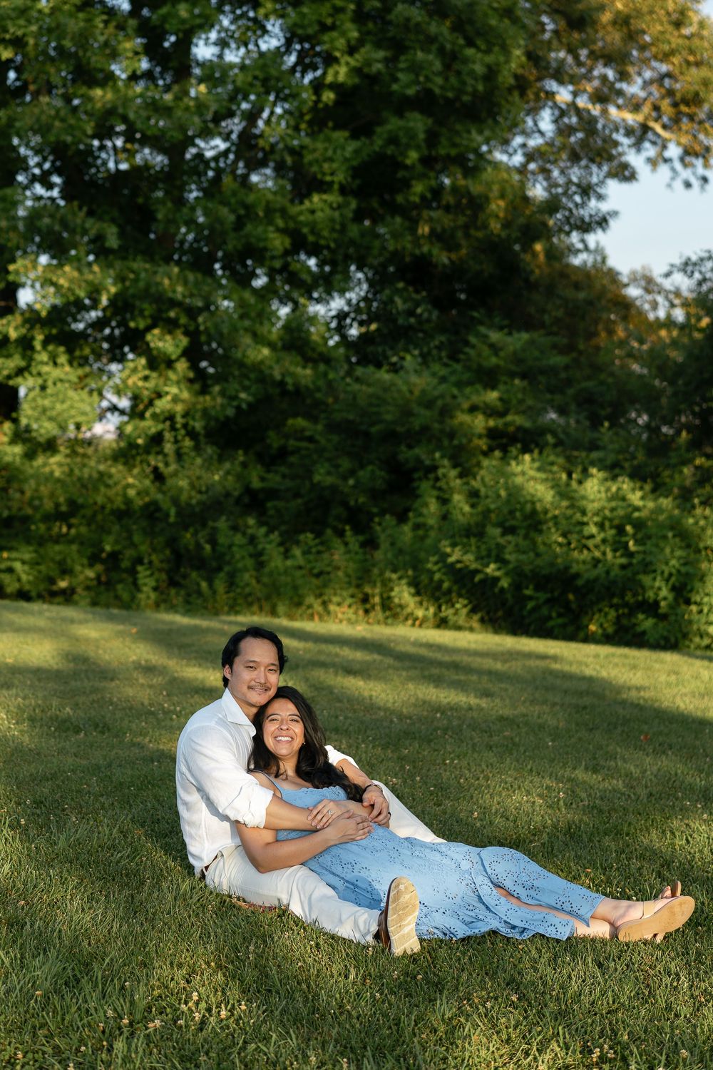 A tender moment captured between a couple sitting together on a green hillside during sunset.