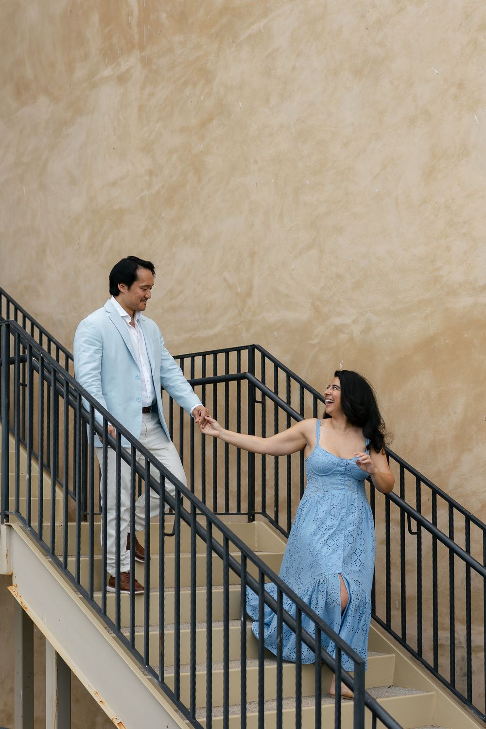 Couple on outdoor concrete staircase with beige wall, person in blue sundress and person in light colored suit.