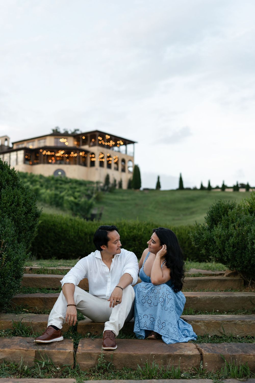 A couple sits on stone steps at a scenic Tuscan villa with cypress trees and string lights at sunset.