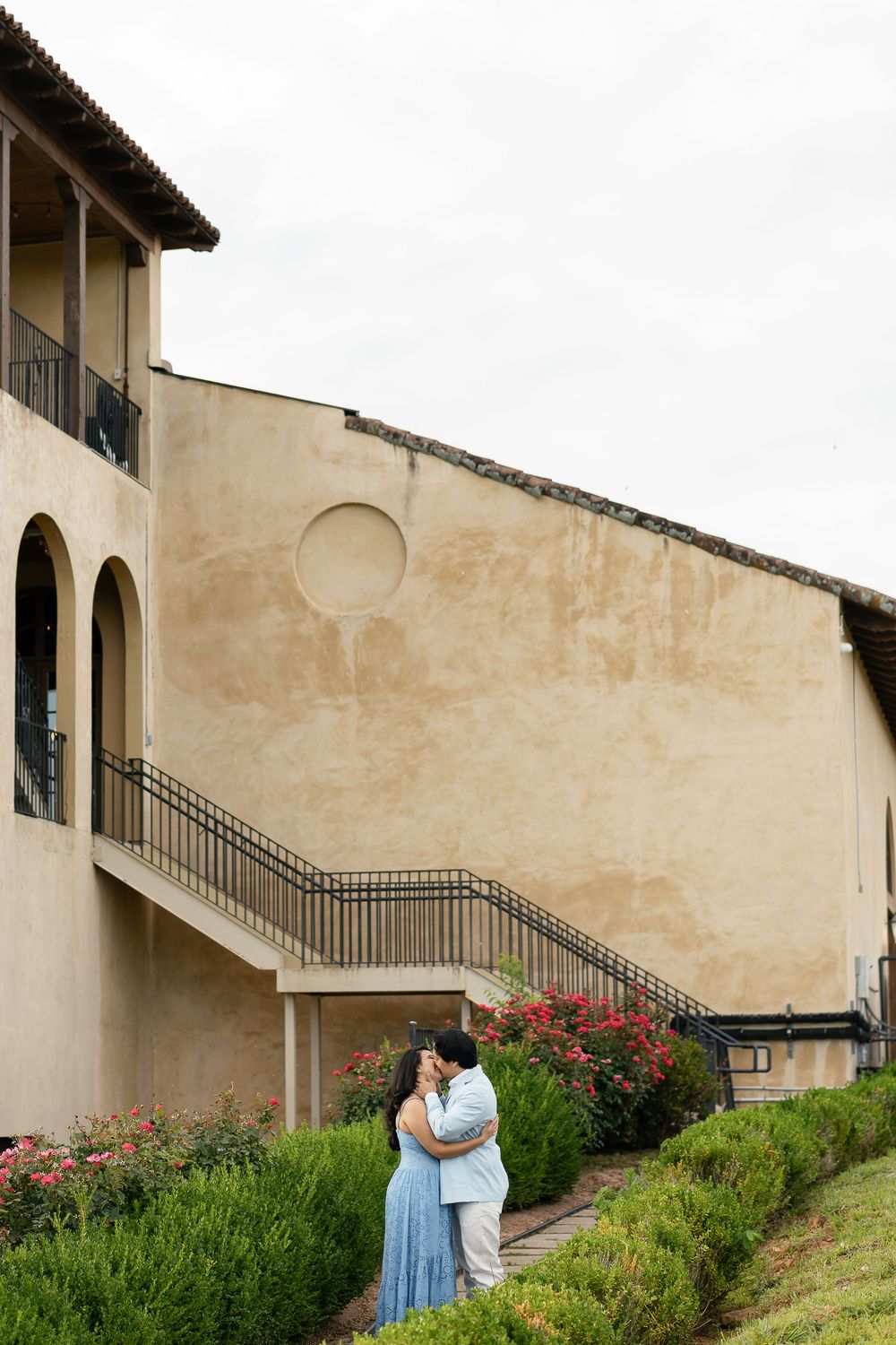 Romantic couple sharing a tender embrace on a landscaped hillside path beside a Mediterranean-style building.