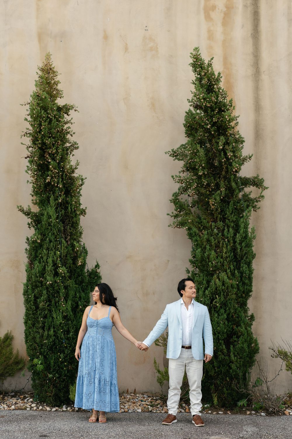 A couple holds hands while standing between two tall cypress trees against a neutral wall.