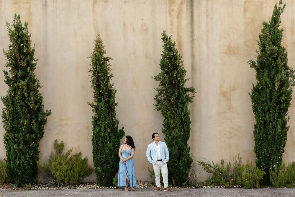 Two people in soft blue outfits stand against a beige wall with tall cypress trees creating vertical lines.