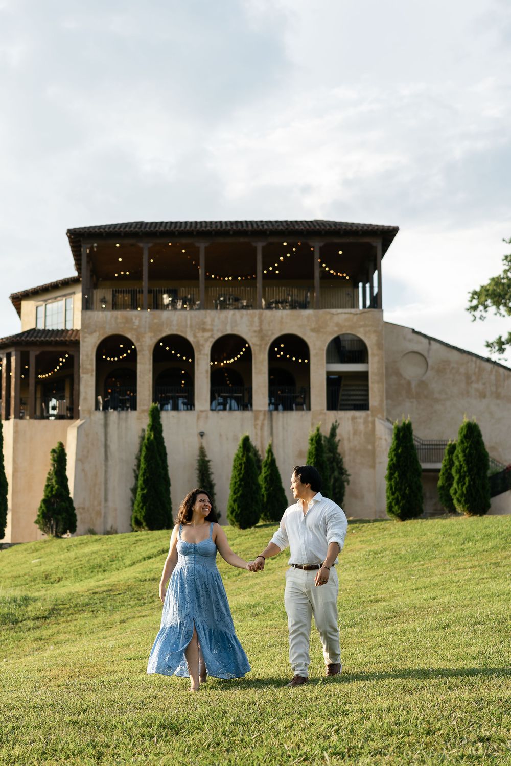 A couple strolls on a grassy hill in front of an elegant venue with string lights and Mediterranean architecture.