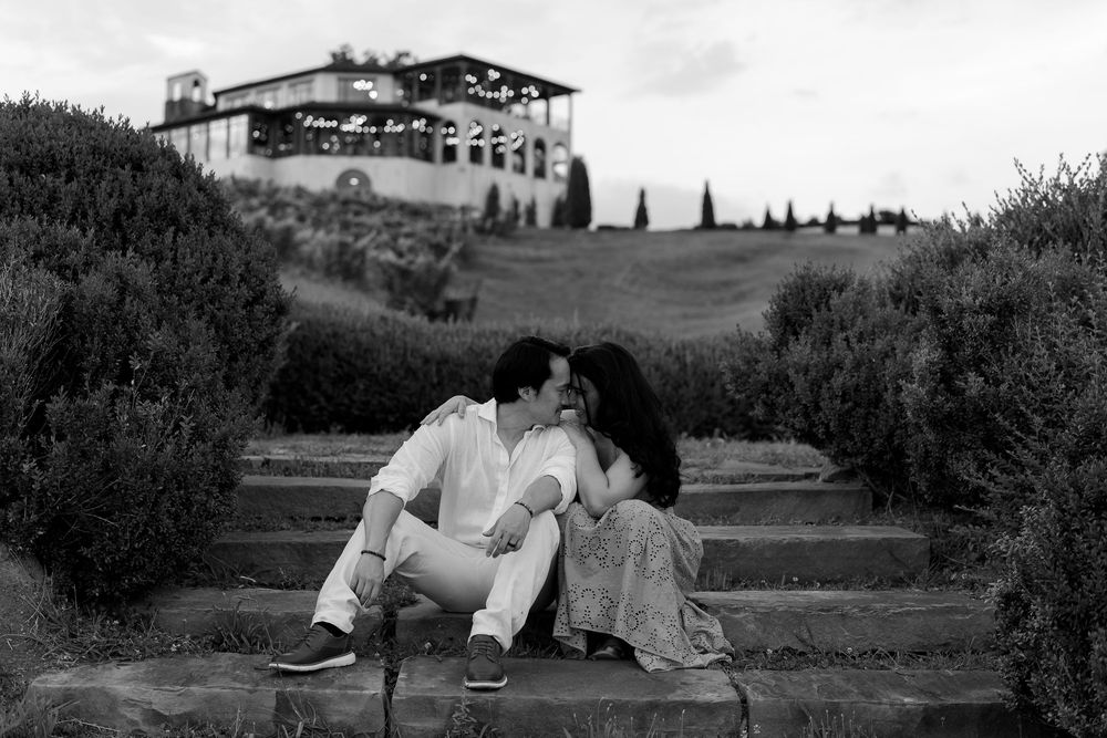 A romantic black and white silhouette of a couple kissing on steps with a villa on a hilltop in the background.