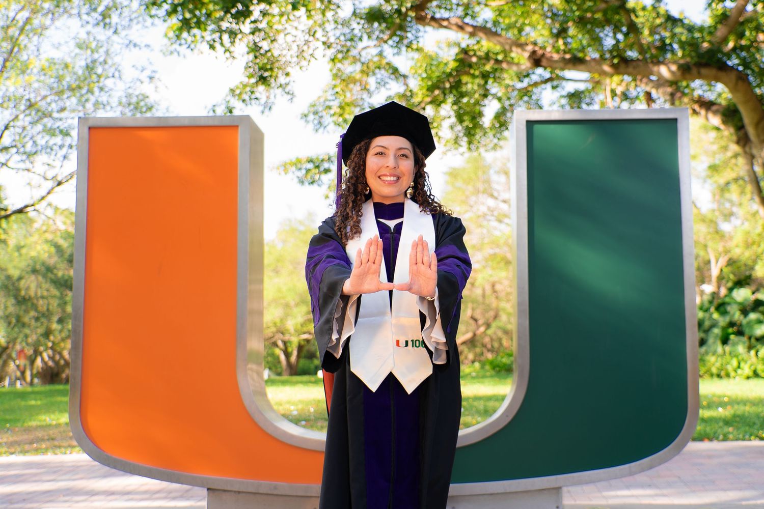 Graduate in cap and gown poses with University of Miami U sculpture making Canes hand sign.