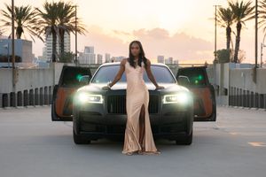 Woman in gold slit gown posing before black Rolls-Royce at sunset with palm trees.