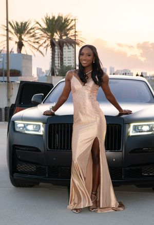 Glamorous woman in gold sequin gown leaning on black Rolls-Royce at golden hour.