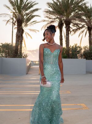 Woman in teal mermaid sequin gown standing among palm trees at sunset.