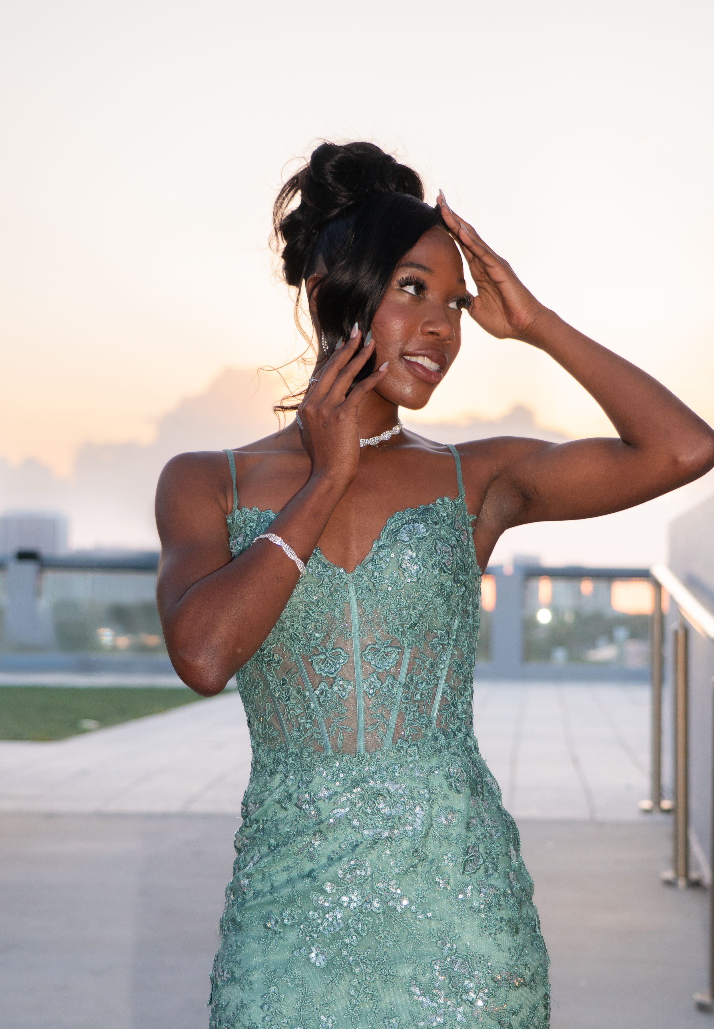 Young woman in green beaded corset gown adjusting hair at rooftop sunset venue.