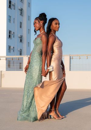 Two women in formal prom gowns, teal and gold, posing back-to-back on rooftop.