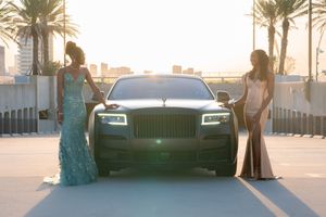 Two women in formal gowns flanking a black Rolls-Royce on a palm-lined boulevard.