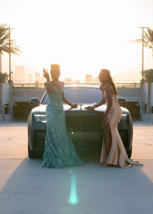 Two elegant women in formal gowns pose in front of a Rolls-Royce at sunset with city skyline backdrop.