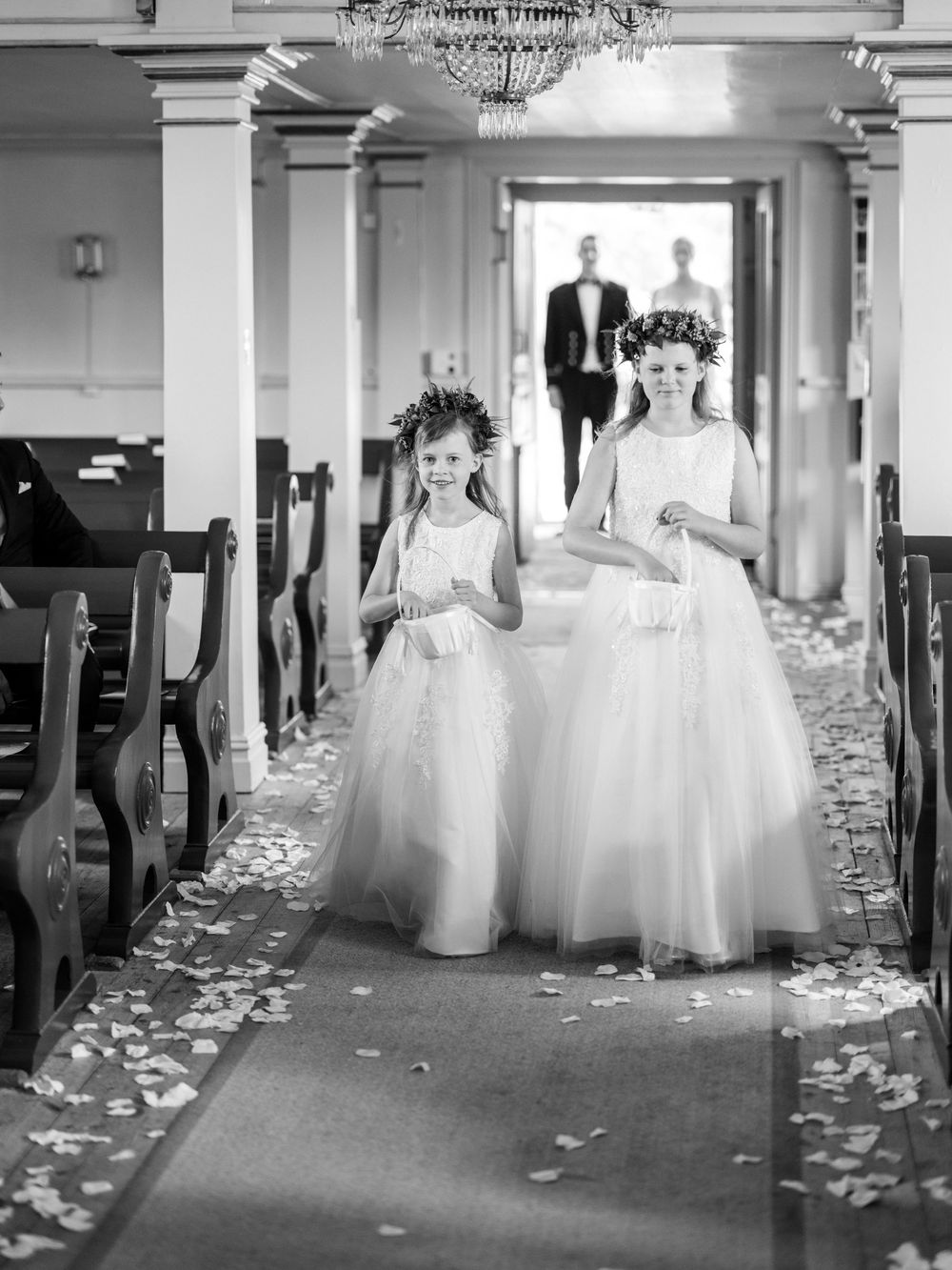 Black and white photo of flower girls in white tulle dresses walking down a rose petal-covered church aisle.