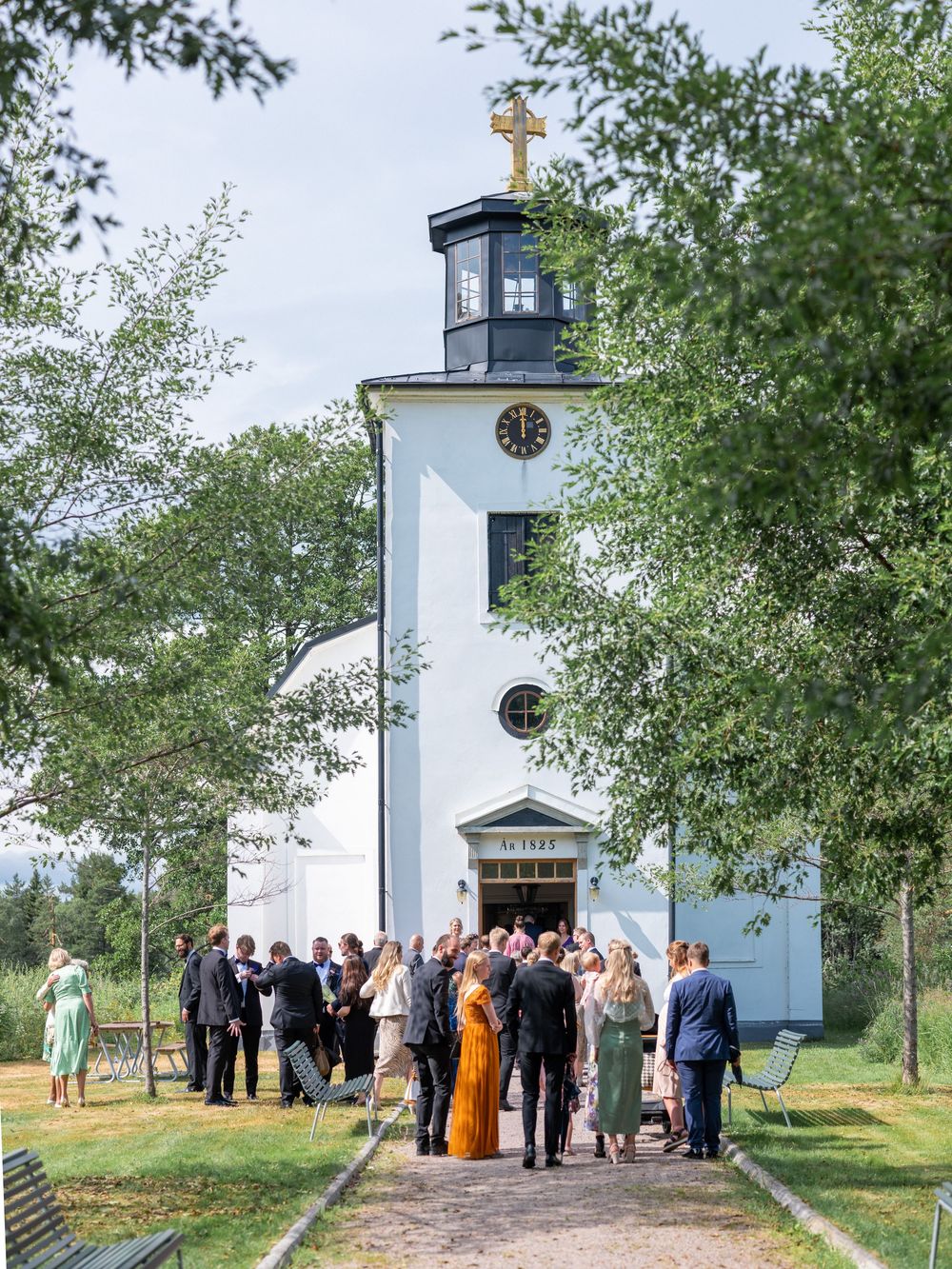 Wedding guests gather outside a white church with a cross and clock tower surrounded by green trees.