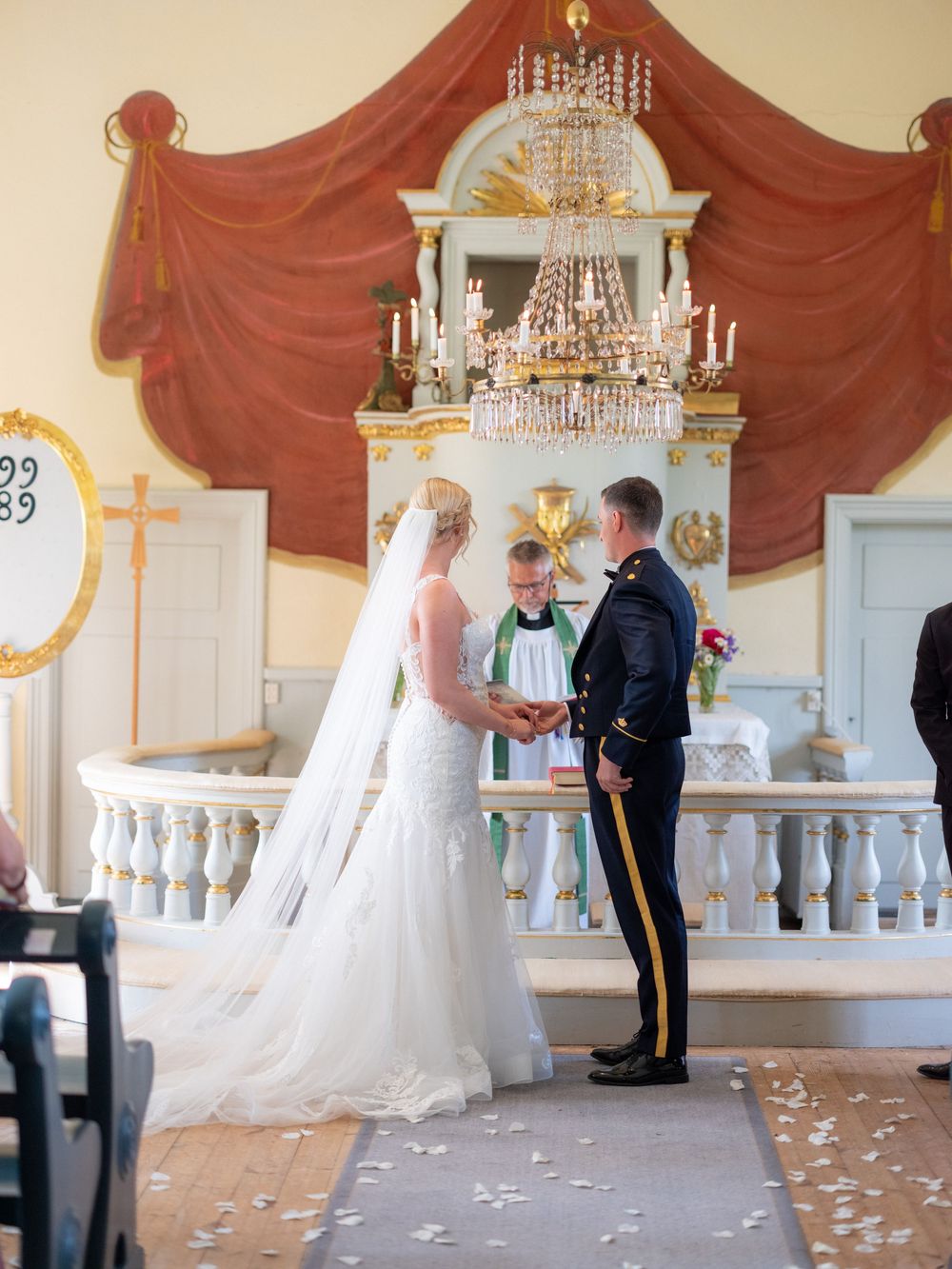 A couple stands at an elegant wedding altar with white pillars, crystal chandelier, and flowing red curtains as rose petals scatter.