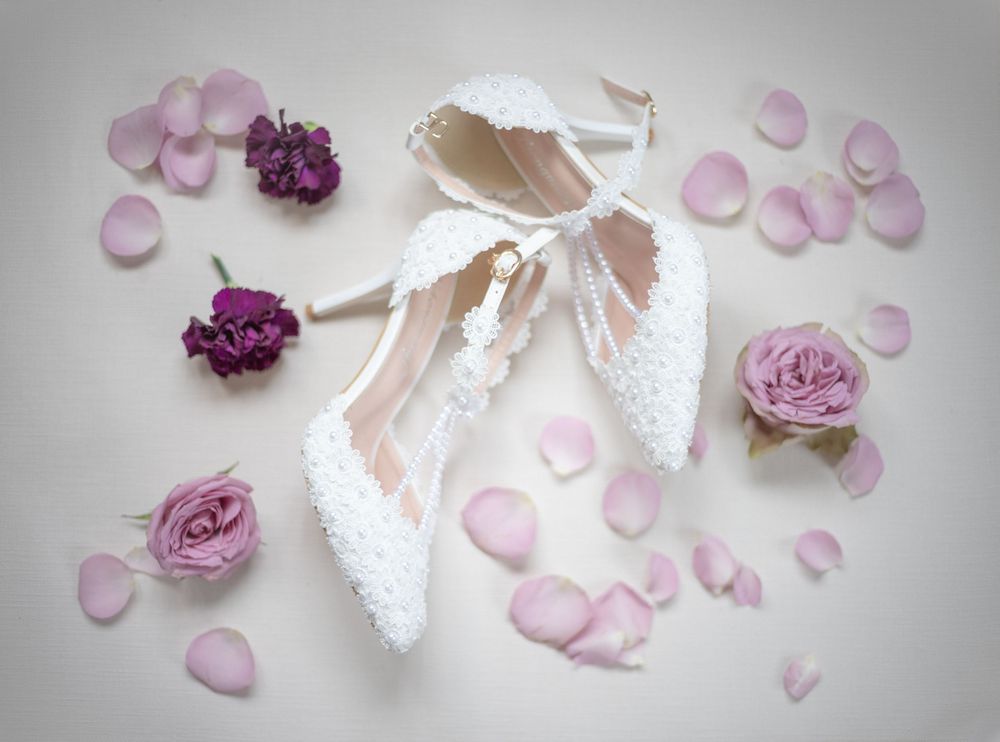 White lace bridal shoes surrounded by scattered purple roses and pink petals on a light gray background.