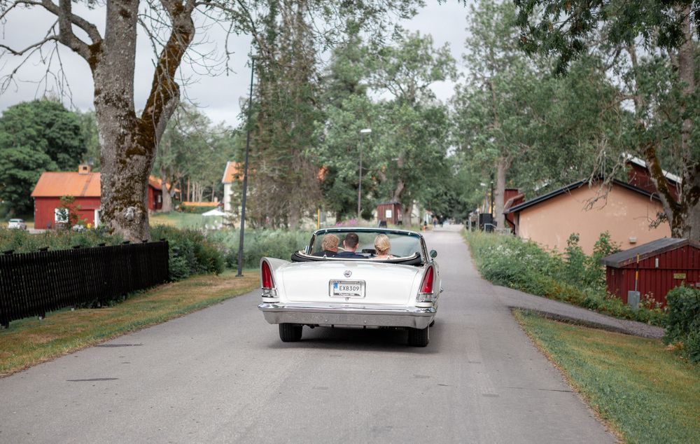 Classic white vintage car drives down tree-lined rural road past red cottages in summer.