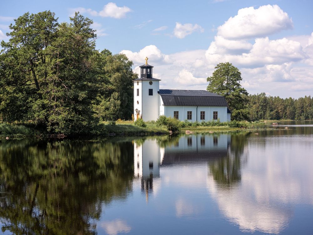 Oslättfors Kyrka - a serene white church with a tall steeple stands by a calm lake, perfectly reflecting in the still waters on a sunny day.