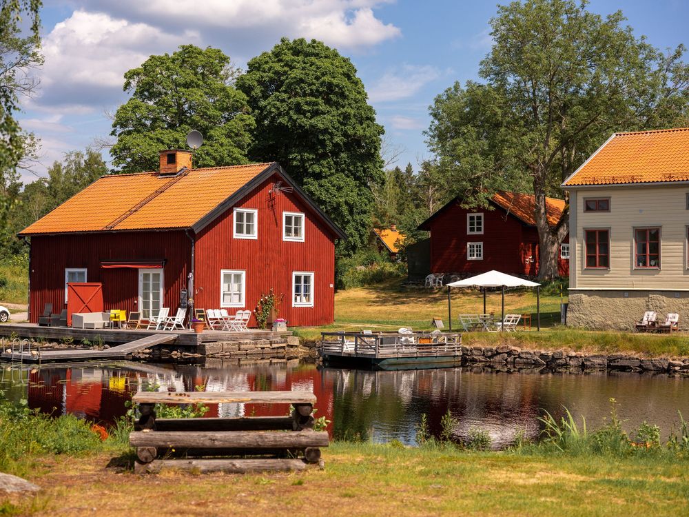 Traditional red Swedish cottages and wooden buildings in Oslättfors line a waterfront on a sunny summer day.