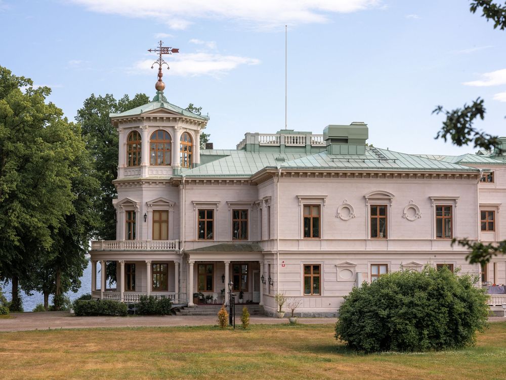 Historic white mansion Engeltofta Hotel and Conference with ornate cupola and green roof sits on a grassy lawn amidst trees on a sunny day.