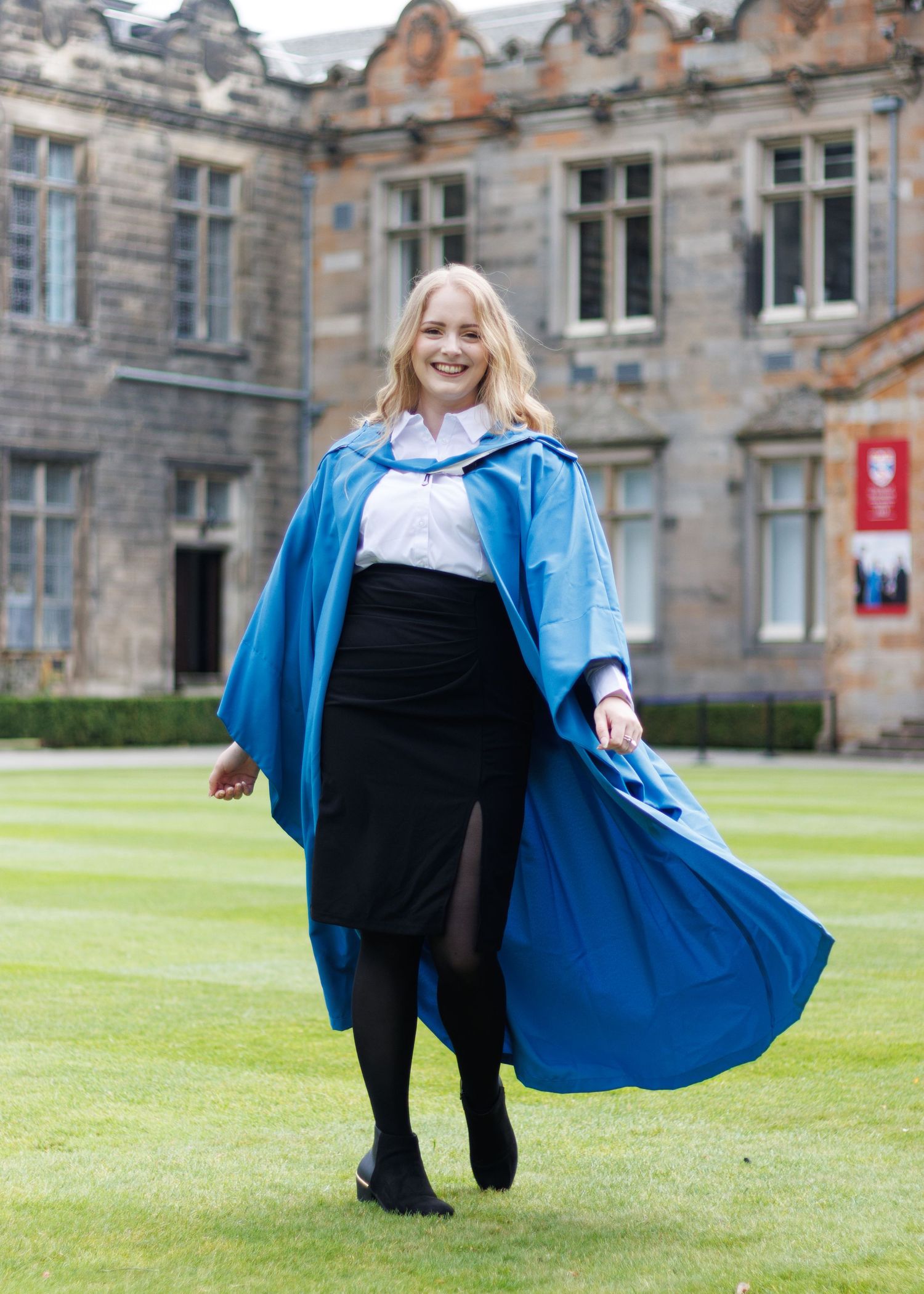 Student wearing blue graduation gown and black skirt smiles while walking on lawn in front of historic stone university building.