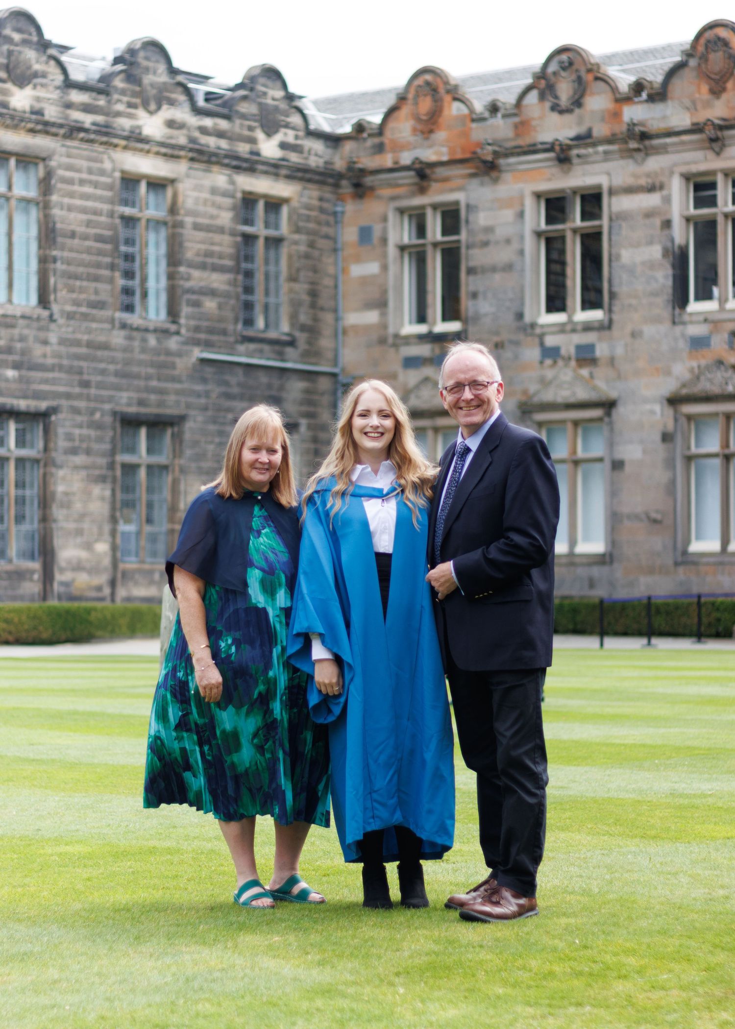 Three people in formal attire pose together on a well-maintained lawn in front of a historic stone building.