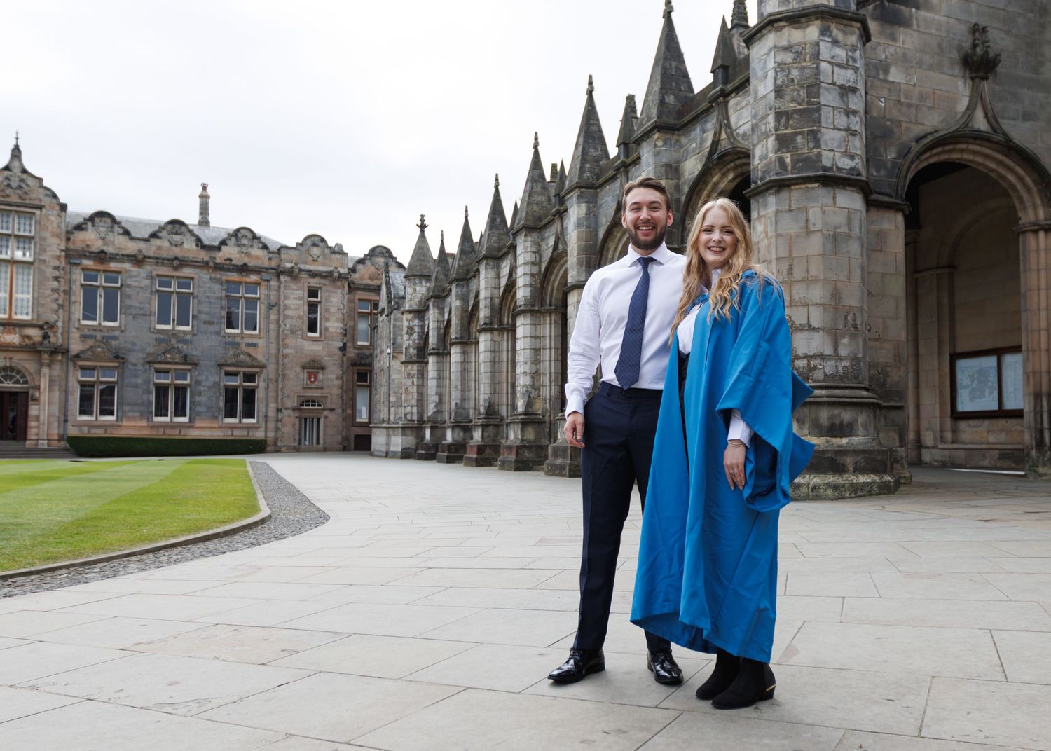 Multiple people in matching blue jackets stand in front of a stone Gothic building on a cloudy day.
