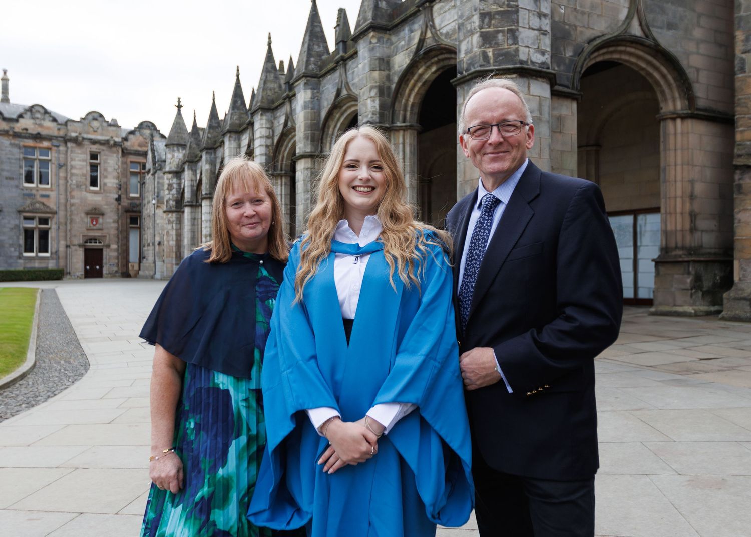 Group photo of graduates wearing blue academic regalia poses with family members outside gothic architecture.