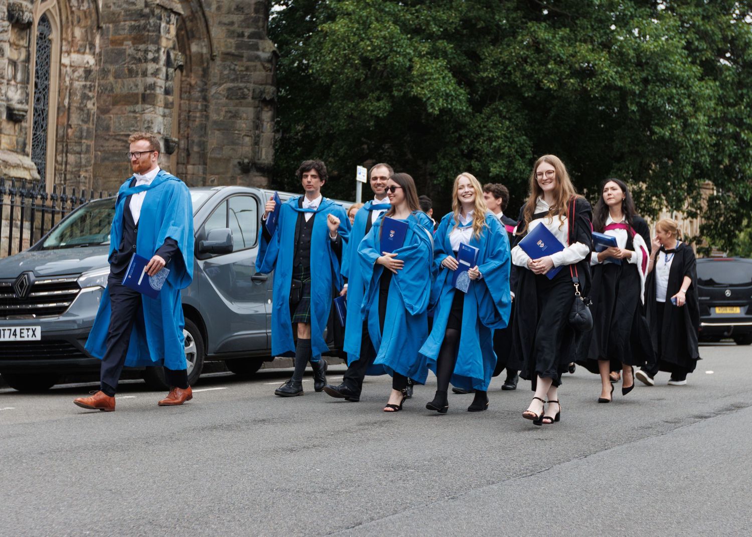 Row of graduates in blue gowns gather outdoors for photos during commencement day celebrations.