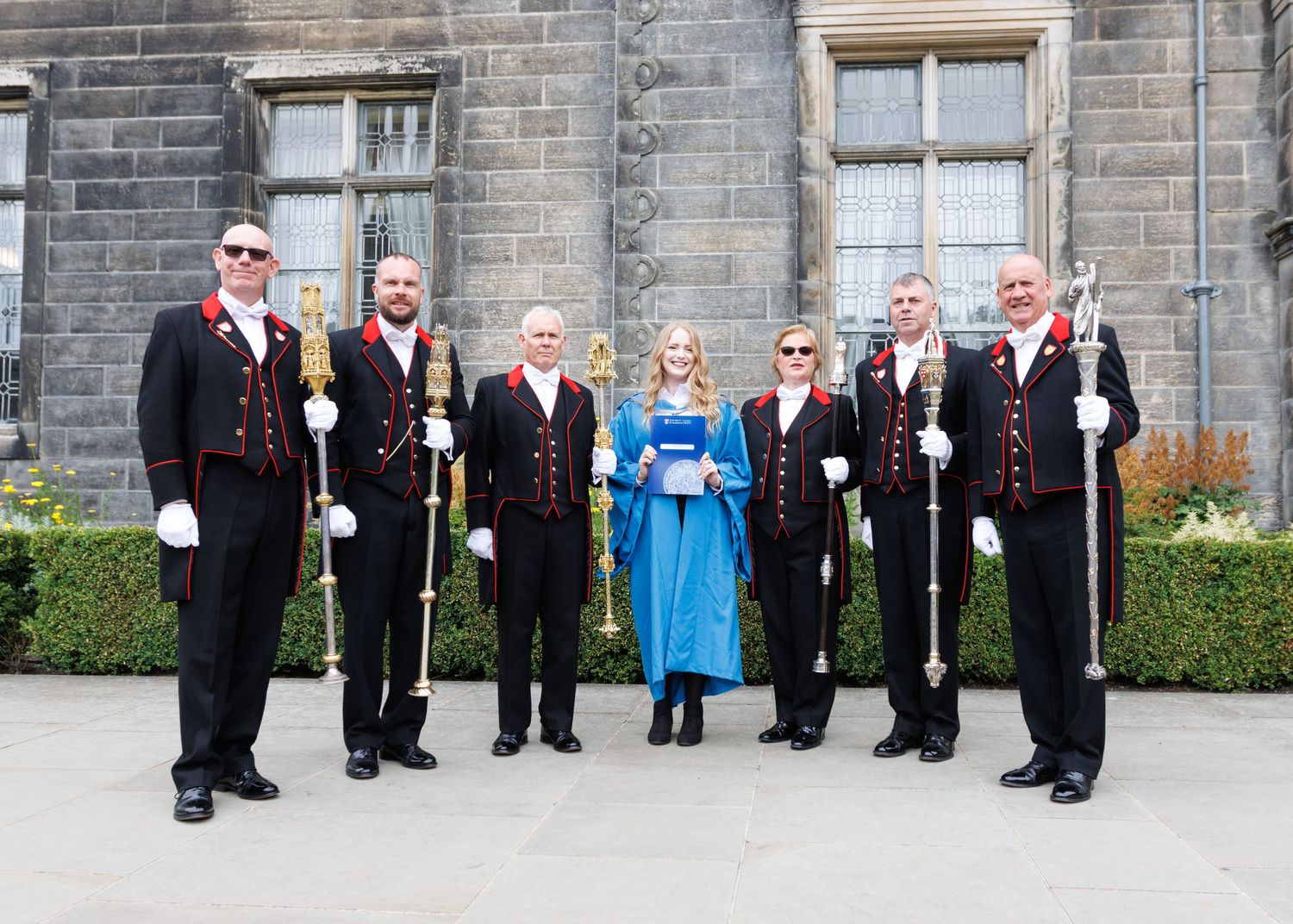 Academic procession of graduates and faculty members lined up along a stone building exterior.