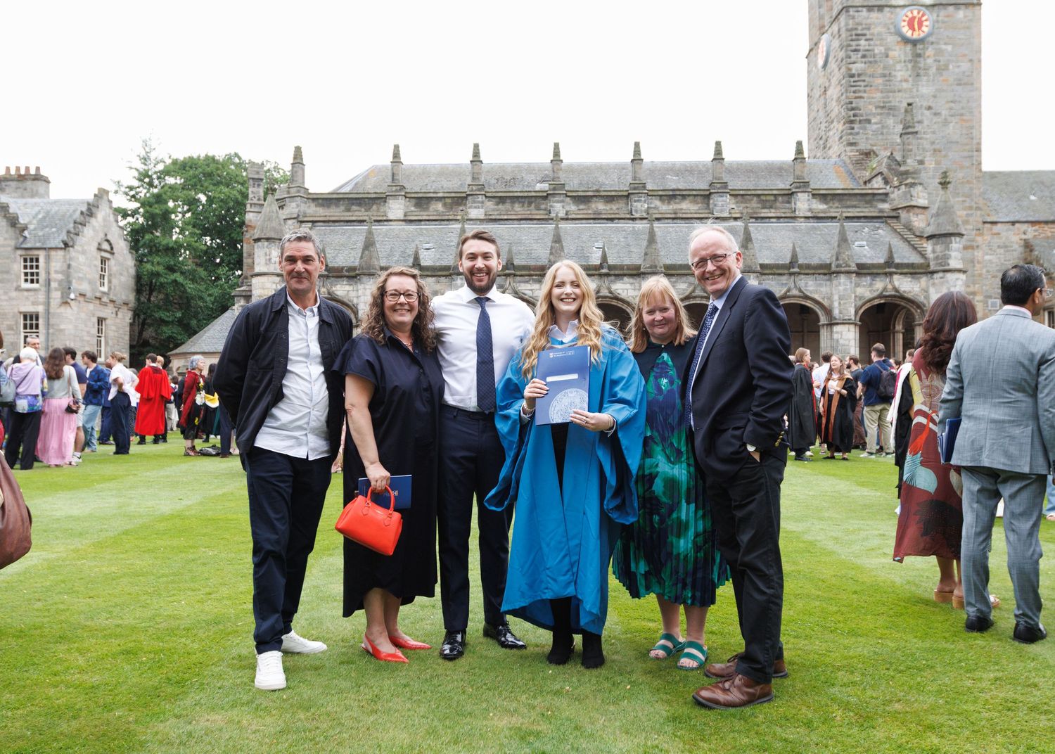 Graduates and guests mingle on a grassy lawn outside historic university buildings during graduation festivities.