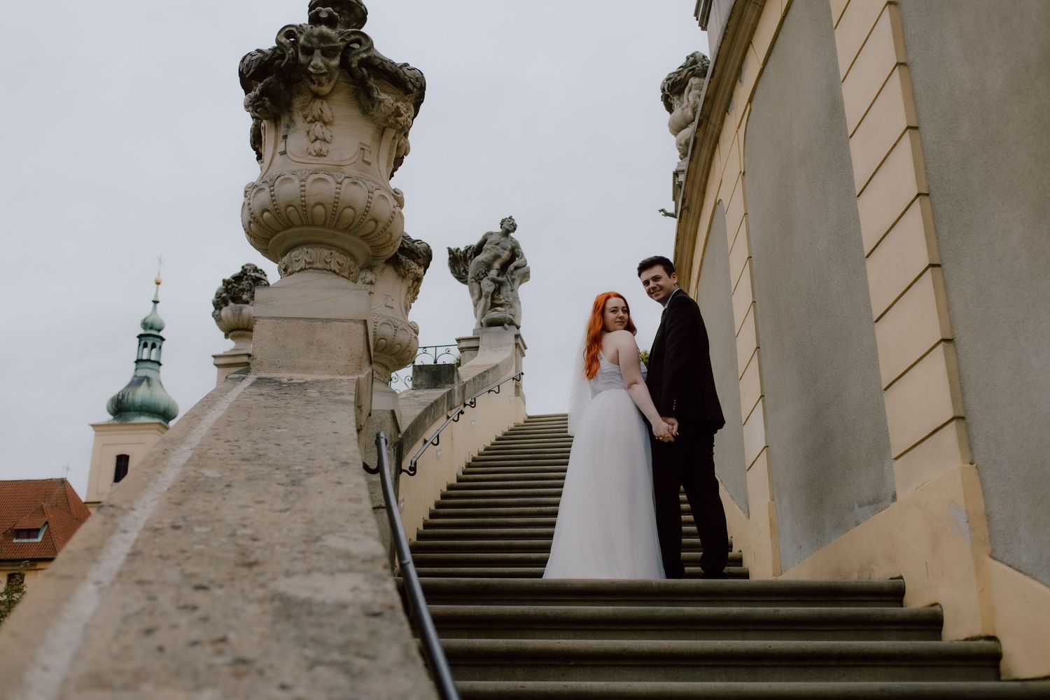 A couple in wedding attire stands on grand stone steps with ornate carved columns and architecture in the background.