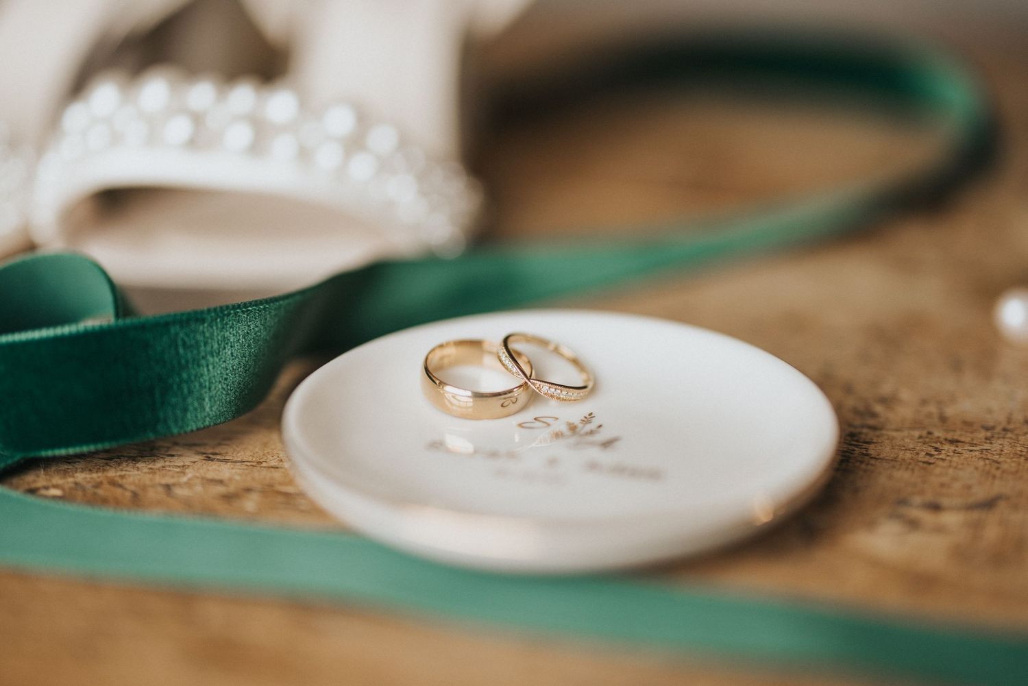 Wedding rings displayed on a white porcelain dish with an emerald green ribbon beside it.