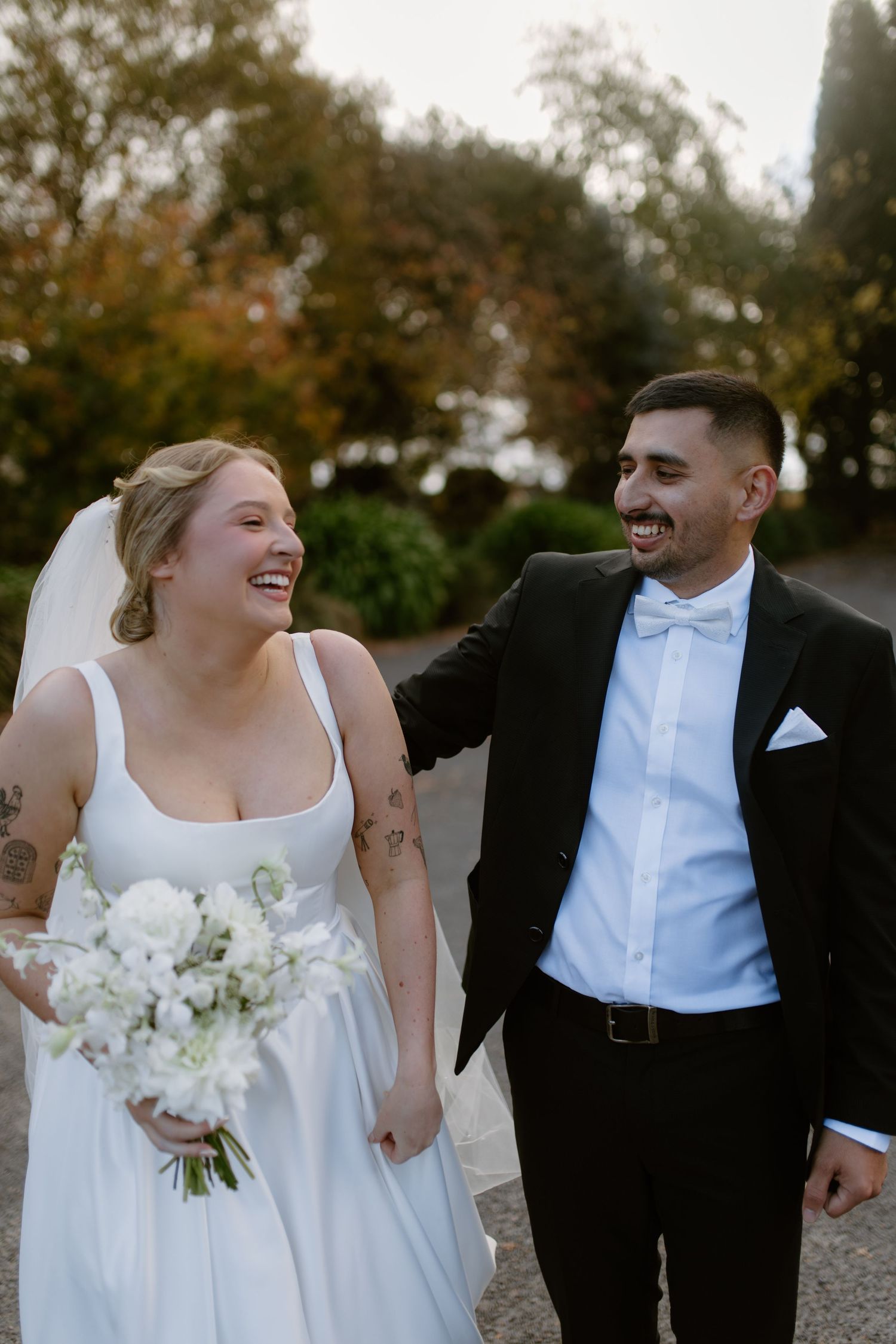 Laughing newlyweds walking together, bride in white gown with bouquet, groom in black tuxedo with bow tie.