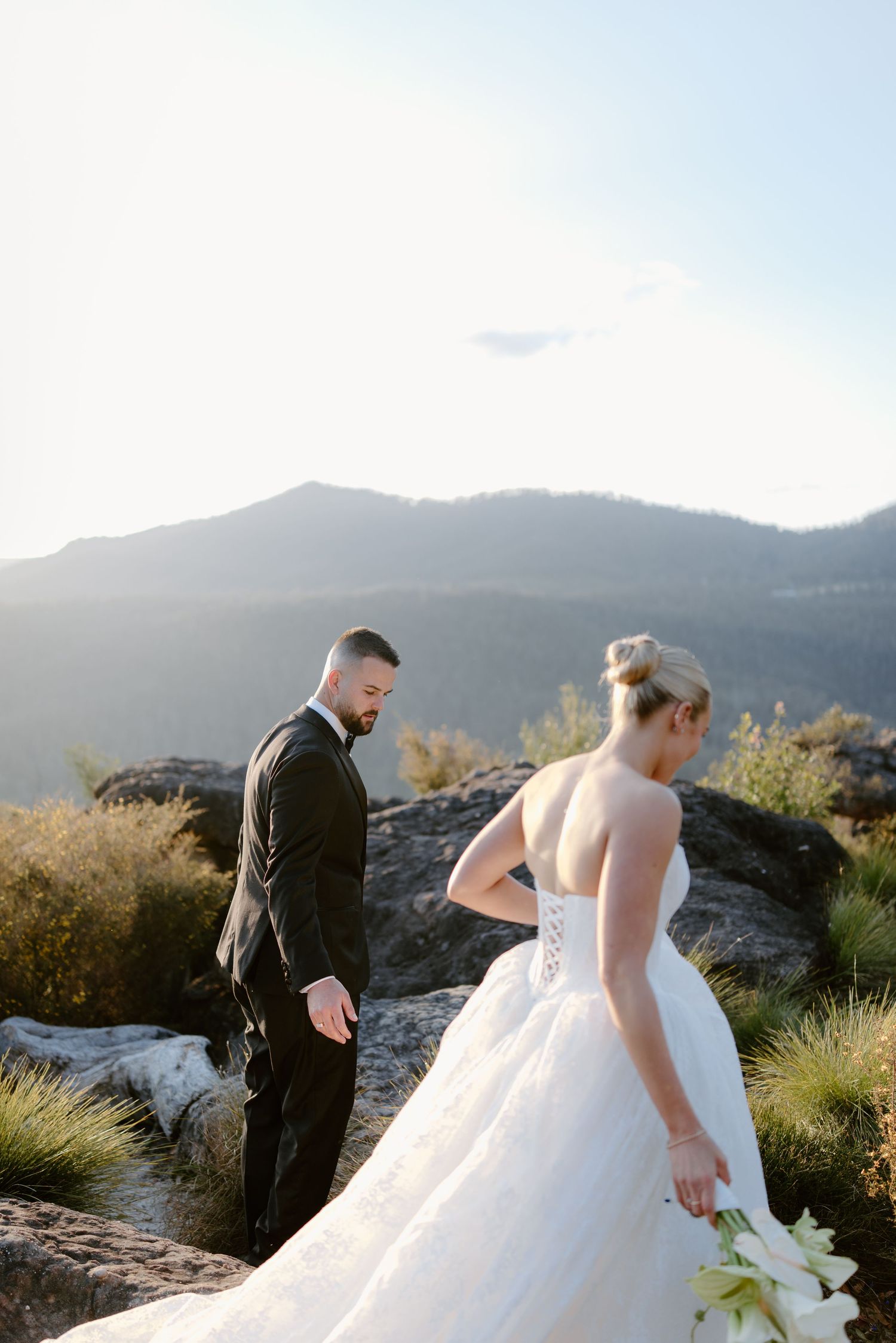 Bride and groom share a romantic moment on a rocky mountain overlook, with stunning misty peaks and golden sunlight behind them.