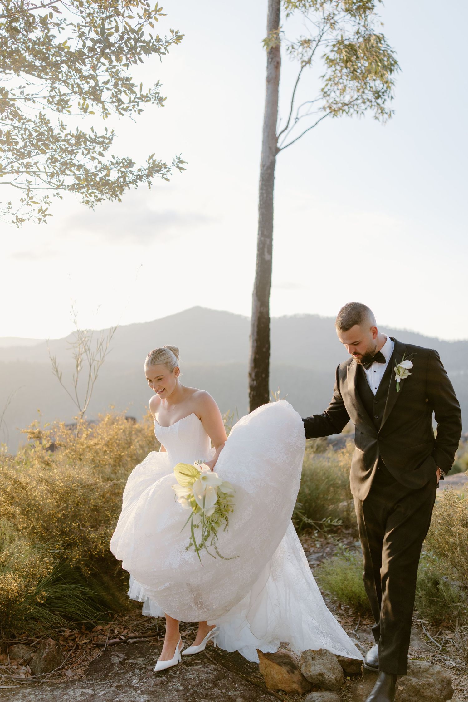 Bride in white gown and groom in black tuxedo walk together on a rocky mountain overlook at golden hour.